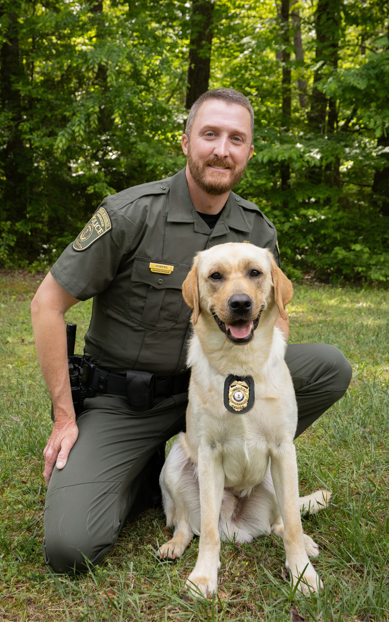 제복을 입은 법 집행관이 목걸이에 배지를 단 개 옆에 무릎을 꿇고 있는 사진입니다.