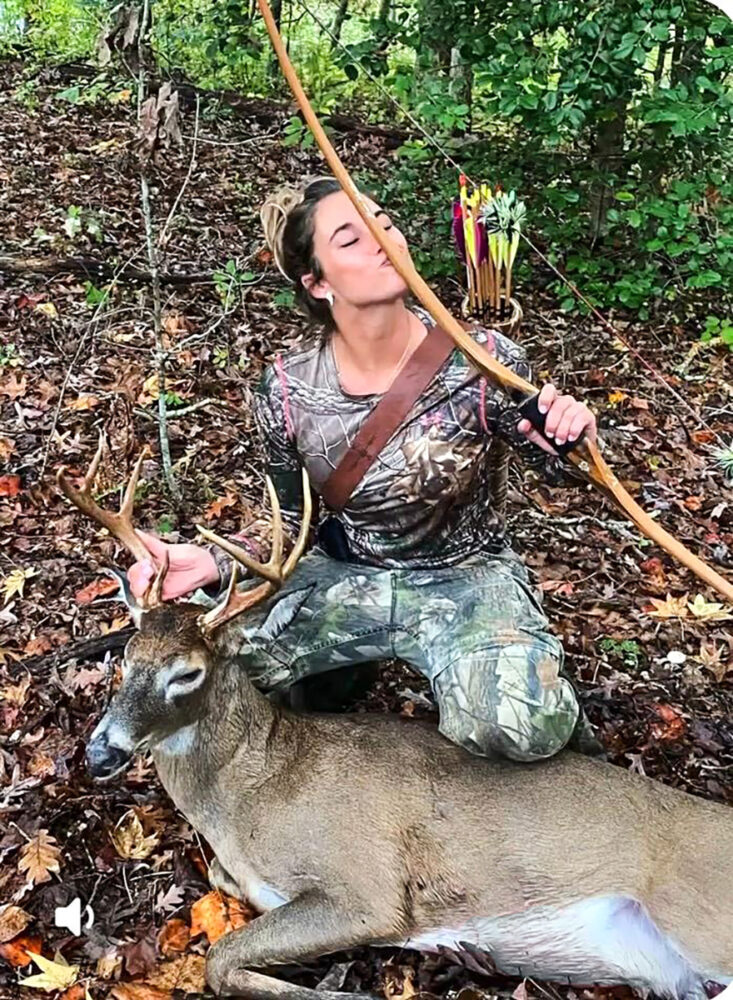 흰꼬리사슴 뒤에 무릎을 꿇고 있는 한 여성의 사진. 그녀는 전통 활을 들고 화살통을 등에 짊어진 채 활에 입맞춤을 하고 있습니다.