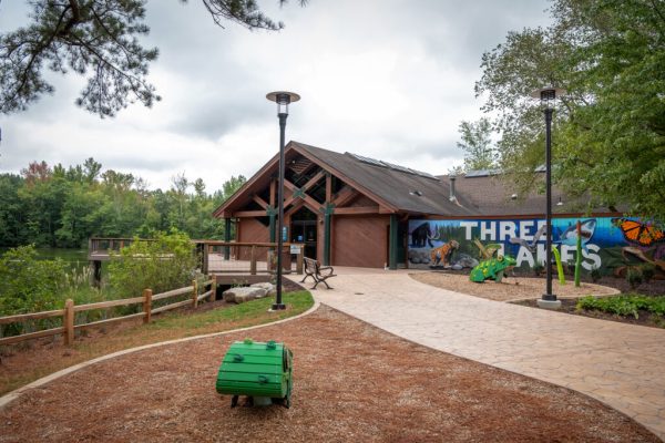 네이처 센터의 정문은 한쪽 벽에 "Three Lakes"라고 쓰인 벽화가 그려진 갈색 건물입니다. 건물 왼쪽에는 호수 중 한 곳을 내려다보는 나무 전망대가 있습니다.